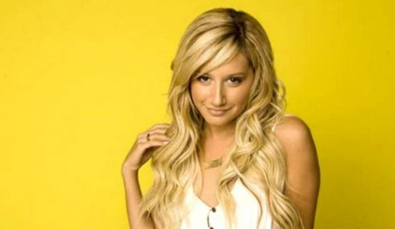 Ashley Tisdale s dlouhými vlnitými vlasy v bílé halence na žlutém podkladu